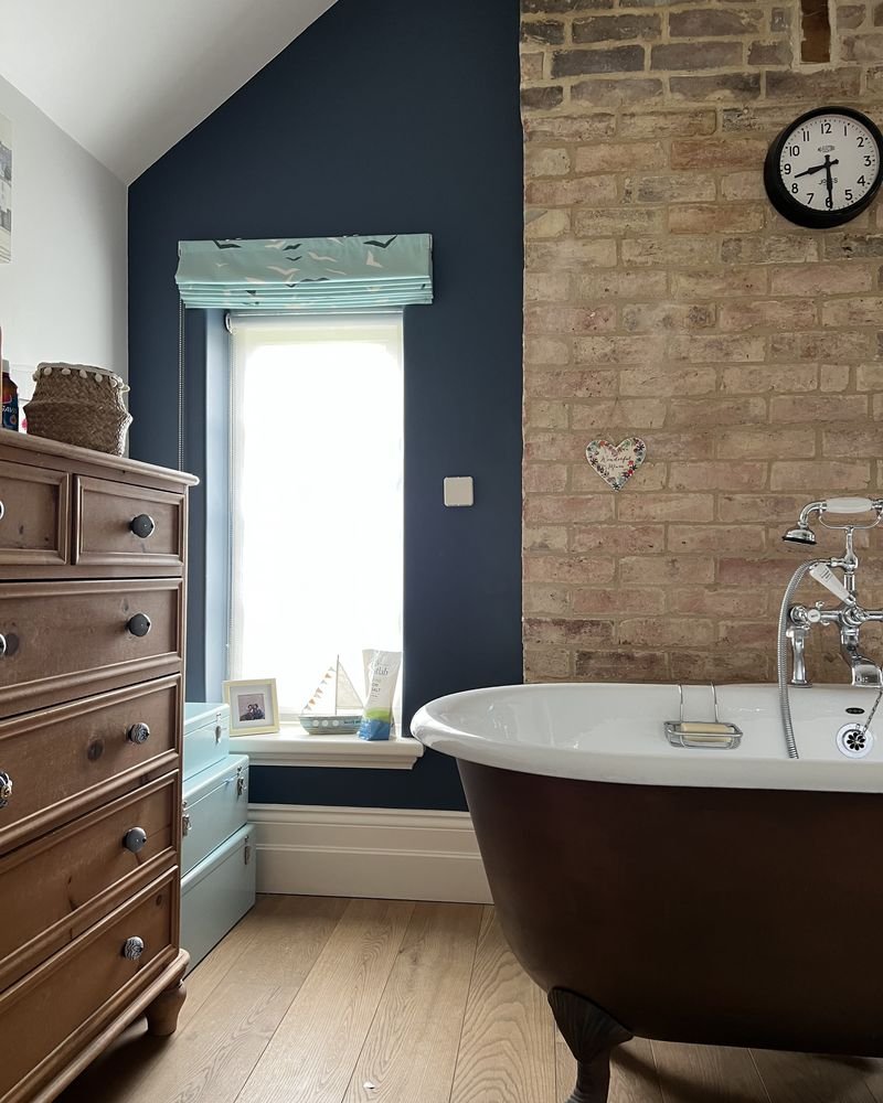 A copper roll-top bath against exposed brick and a navy wall